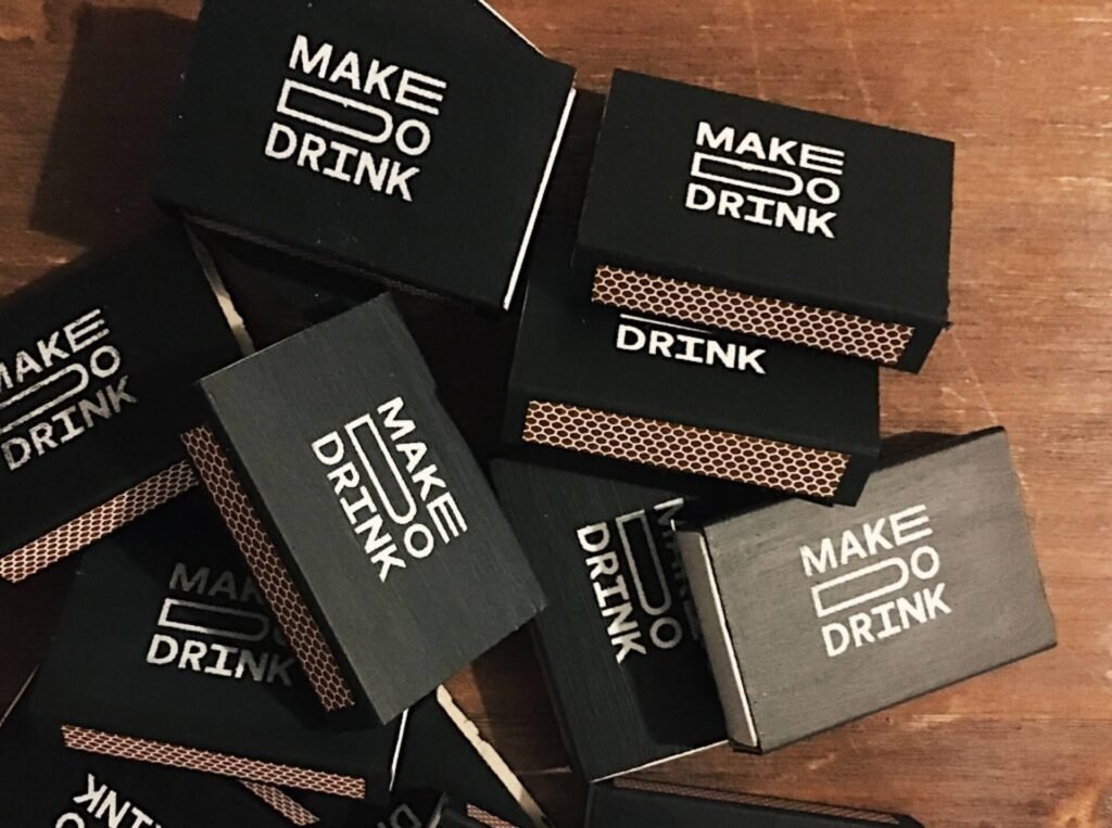 black matchboxes with the make do drink logo on a wooden table